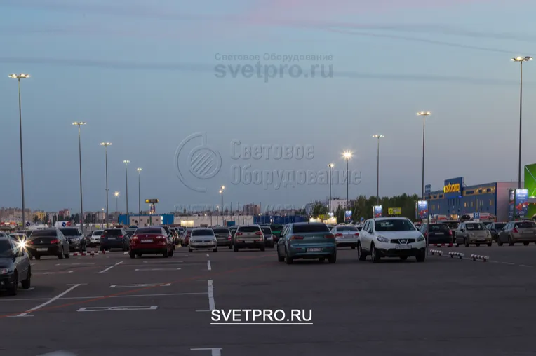 На изображении приведен пример использования опор ОГК с высотой в 16 метров для освещения парковки перед крупным торговым центром. На верхушках установлены многогранные кронштейны для крепления прожекторов с направленным световым лучом.