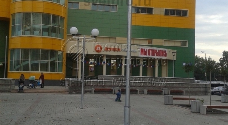 На одной опоре из стальных труб можно установить несколько светильников, как это видно на изображении. Два СП закреплены на двух выносных консолях, направленных в противоположные стороны. За счет этого освещается большее пространство площади перед торговым центром и сокращается число опор. 