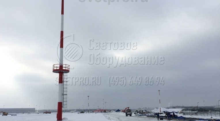 На изображении представлена мачта для освещения аэродрома, которая имеет неподвижную круглую площадку в нижней части и раму спускаемого типа вверху. Для обеспечения большей безопасности и соблюдения международных регламентов безопасности полетов корпус окрашен в красные и белые полосы.